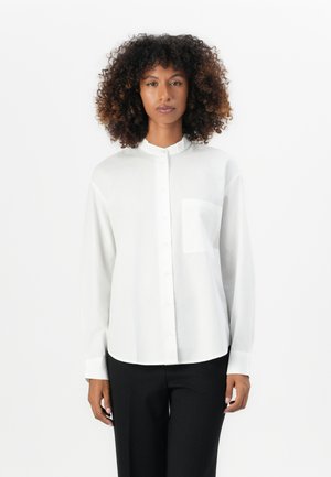 Camicia bianca a maniche lunghe con colletto alla coreana, chiusura a bottoni e taschino sul lato sinistro del petto. Tessuto morbido e vestibilità comoda, abbinata a pantaloni neri.