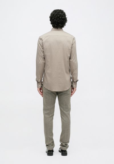 Light brown button-up shirt with long sleeves, back yoke, and curved hem, paired with grey textured trousers and black shoes.
