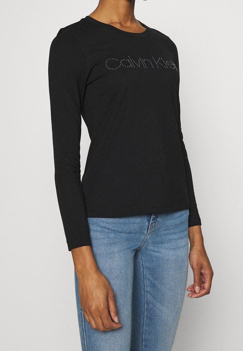 T-shirt noir à manches longues en tissu doux, avec un col arrondi et un logo en strass sur le devant. Associé à un jean bleu clair.