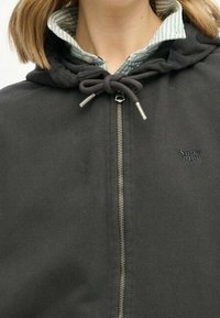 Chaqueta negra con cremallera y capucha ajustable, que presenta un cuello interior a rayas verdes y blancas y un pequeño logo de Superdry.