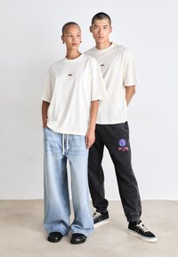 To personer står side om side. Den ene har en løs cremefarvet t-shirt på med et lille logo og baggy lyseblå jeans. Den anden har en sort t-shirt og mørke sweatpants med grafik. Begge har casual sorte sneakers på.