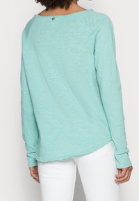 Helles türkisfarbenes Langarmshirt aus weichem Stoff, mit einem runden Halsausschnitt und einem kleinen Knopfdetail auf der Rückseite.