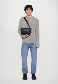 Schwarze, rechteckige Tasche mit Reißverschluss und silbernen Hardware, gehalten von einem Modell in einem hellgrauen Rippensweater und blauen Jeans.