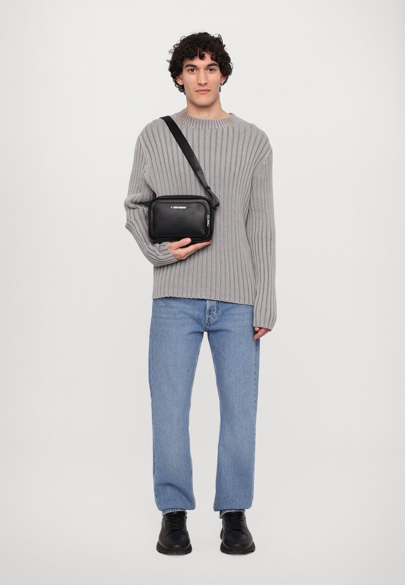 Schwarze, rechteckige Tasche mit Reißverschluss und silbernen Hardware, gehalten von einem Modell in einem hellgrauen Rippensweater und blauen Jeans.