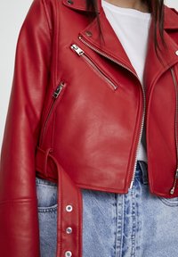 Gros plan d'une personne portant une veste de motard en cuir rouge avec des fermetures éclair et des boutons argentés, par-dessus une chemise blanche et un jean bleu.