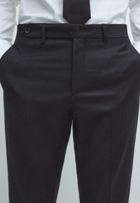 Zwarte formele broek met een gladde textuur, platte voorkant, zijzakken en een knoopsluiting. Gecombineerd met een wit overhemd en een stropdas.