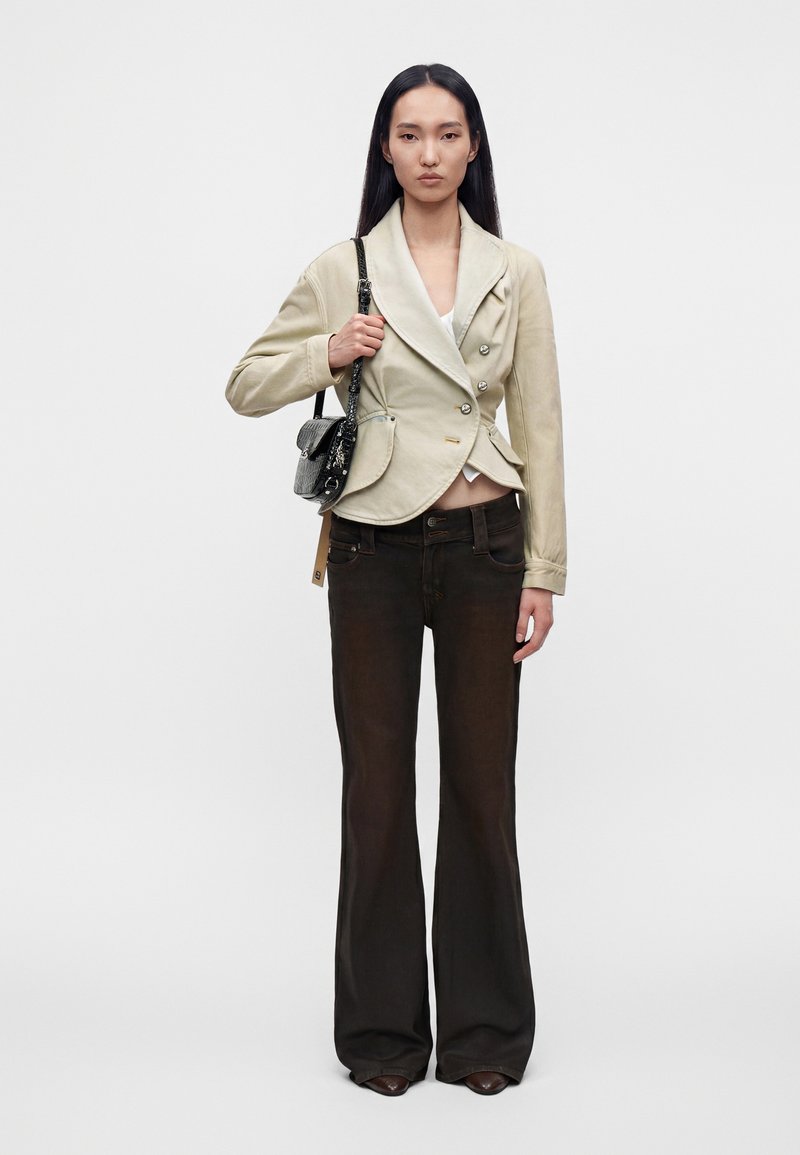 Woman with long black hair wears a beige double-breasted jacket, dark flared pants, brown shoes, and carries a small black handbag on her shoulder.