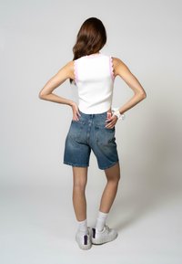 Haut sans manches blanc avec des bords festonnés roses, associé à un short en denim bleu clair. Le modèle porte des baskets blanches avec des accents violets.