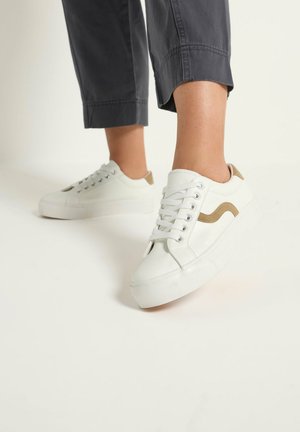 Persona che indossa sneakers bianche con dettagli beige e pantaloni neri corti, in piedi su un pavimento bianco davanti a uno sfondo semplice.