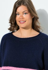 Jonge vrouw met schouderlang bruin haar, gekleed in een blauwe gebreide trui met een roze streep, die glimlacht tegen een witte achtergrond.
