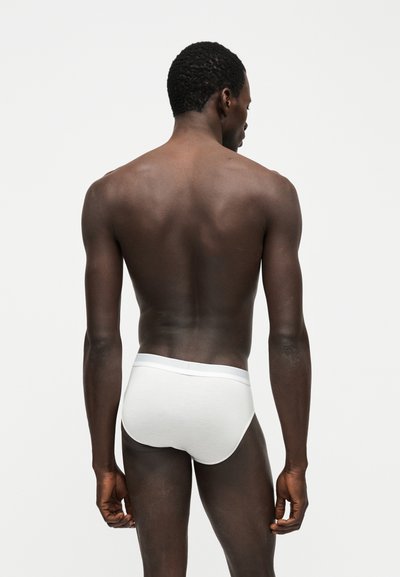 Άνδρας που στέκεται με την πλάτη προς τα έξω φορώντας απλά λευκά εσώρουχα μπροστά σε απλό ανοιχτό γκρι φόντο.