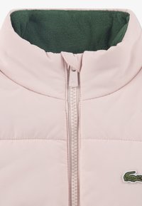 Svetlo roza puhasta jakna z zeleno flisasto podlogo in zadrgo na sprednji strani. Na levi strani prsnega koša je majhen zeleni vezen logotip.