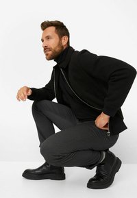 Zwarte zip-up jack gedragen over een zwarte coltrui, gecombineerd met grijs gemusterde broek en zwarte enkellaarsjes. De outfit heeft een moderne, op maat gemaakte uitstraling.