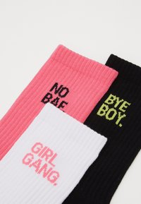 Trīs pāri ribotajām zeķēm: rozā ar uzrakstu "NO BAE," baltas ar uzrakstu "GIRL GANG" un melnas ar uzrakstu "BYE BOY," ar kontrastējošām teksta krāsām.