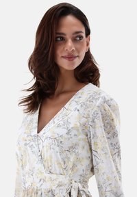 Weißes Blumenmusterkleid mit kurzen Puffärmeln, das gelbe und grüne Blumenmuster auf einem leichten Stoff zeigt. V-Ausschnitt mit Bindeband.