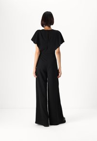 Svart jumpsuit med vide, flagrende ermer og en avslappet passform, med en glatt tekstur og en lukking bak i midten.