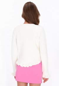 Donna con lunghi capelli castani che indossa un maglione bianco a coste con bordi arricciati e una gonna corta di un vivido colore rosa, rivolta di spalle rispetto alla fotocamera.