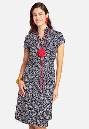 Woman wearing a navy dress with white floral pattern, red beaded necklace with flower, red earrings, and gold bangles, smiling with hand on hip.