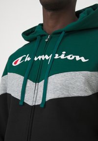 Sudadera con cremallera verde y gris, con fondo negro, que presenta un logo blanco en el pecho, cordones y un diseño de bloque de color en forma de chevron.