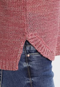 Pull en maille rose clair avec un motif texturé et des accents de fil argenté, doté d'une fente sur le côté et de bords côtelés, associé à un jean en denim foncé.
