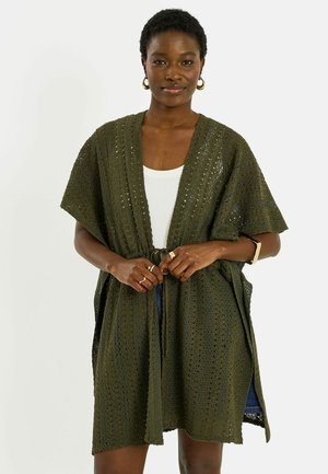 Mujer con cárdigan corto de punto verde oscuro sobre una camiseta blanca y una falda azul, complementada con pendientes de aro dorados y anillos.