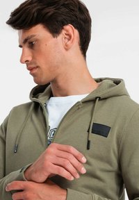 Sudadera con cremallera de color verde oliva, con cordón y un parche de logo negro, que presenta una textura suave y un diseño ajustado, llevada sobre una camisa blanca.