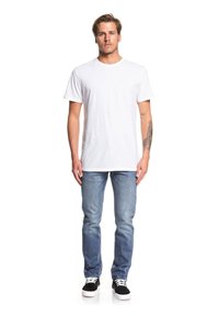 T-shirt bianco in cotone, dalla vestibilità relaxed, abbinato a jeans azzurri chiari. Sneakers nere con suole bianche. Figura maschile in piedi con tatuaggi visibili.