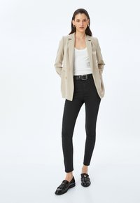 Beige dubbelknäppt kavaj över en vit tanktop, tillsammans med svarta slim fit jeans och svarta loafers med fransdetaljer.