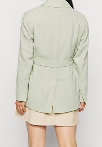 Ljusgrön kavaj i slätt tyg med bälte i midjan, långa ärmar och öppning bak. Bärs över en beige manchesterkjol.