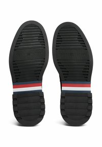 Soli delle scarpe in gomma nera con motivi testurizzati, caratterizzati da una banda accentata a strisce in rosso, bianco e blu, che mostra il nome del marchio "Tommy Hilfiger."