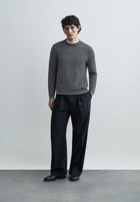 Gray patterned sweater with a round neckline paired with loose black trousers; model stands on a smooth concrete floor against a blank wall.