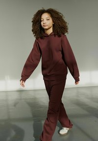 Bordeauxfarbener Kapuzenpullover mit Logo, kombiniert mit passenden bordeauxfarbenen Sweatpants. Glatter Stoff, lockere Passform und weiße Sneakers.