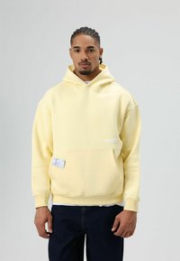 Lichtgele hoodie met een voorzak, geribde manchetten en een label. Het materiaal is zacht, met een losse pasvorm en zonder patronen.