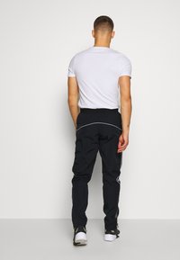 Man med vit kortärmad skjorta, svarta träningsbyxor med reflekterande detaljer och svart-vita sneakers, stående vänd mot en tom vägg.