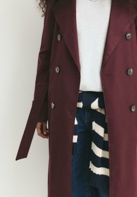 Personne portant un manteau double boutonnage bordeaux par-dessus un pull blanc, avec un pull en tricot rayé bleu et blanc noué autour de la taille.