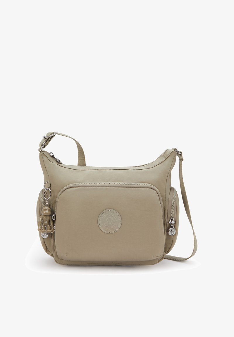 Beige schoudertas met een voorvak, verstelbare riem, zijritsen en een kleine sleutelhanger in de vorm van een aapje bevestigd aan een ritssluiting.