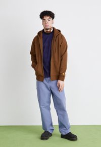 Sudadera marrón con cremallera y bolsillos frontales, llevada sobre una camiseta de rayas oscuras. Unos pantalones azul claro y unos zapatos negros sin cordones completan el conjunto.