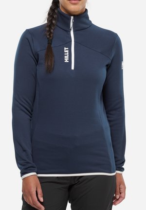 Femme portant un haut bleu marine à manches longues avec fermeture éclair et logo "Millet", associé à un pantalon noir, sur un fond blanc uni.