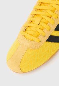 Κίτρινα αθλητικά παπούτσια με υφαντό ύφασμα στο πάνω μέρος και σουέντ λεπτομέρειες· διαθέτουν μαύρες ρίγες και φωτεινά κίτρινα κορδόνια. Ορατή ελαστική σόλα.