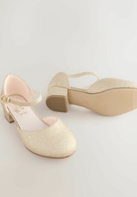 Guldglitrende Mary Jane-sko med lave blokhæle og dekorative spænder på en enkel lys baggrund.