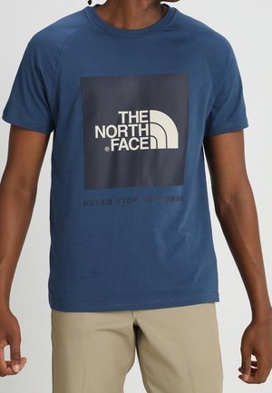 Osoba ubrana w niebieską koszulkę The North Face z logo i hasłem „Nigdy nie przestawaj odkrywać”, w połączeniu z beżowymi spodniami.