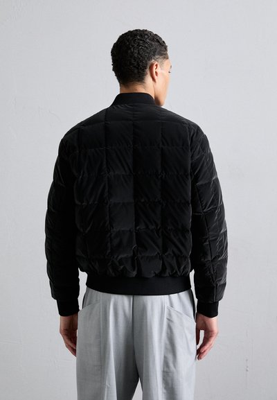 Sort pufferjakke med quiltet mønster, ribbede manchetter og krave. Båret med lysegrå bukser, set fra bagpå. Blødt, polstret materiale.