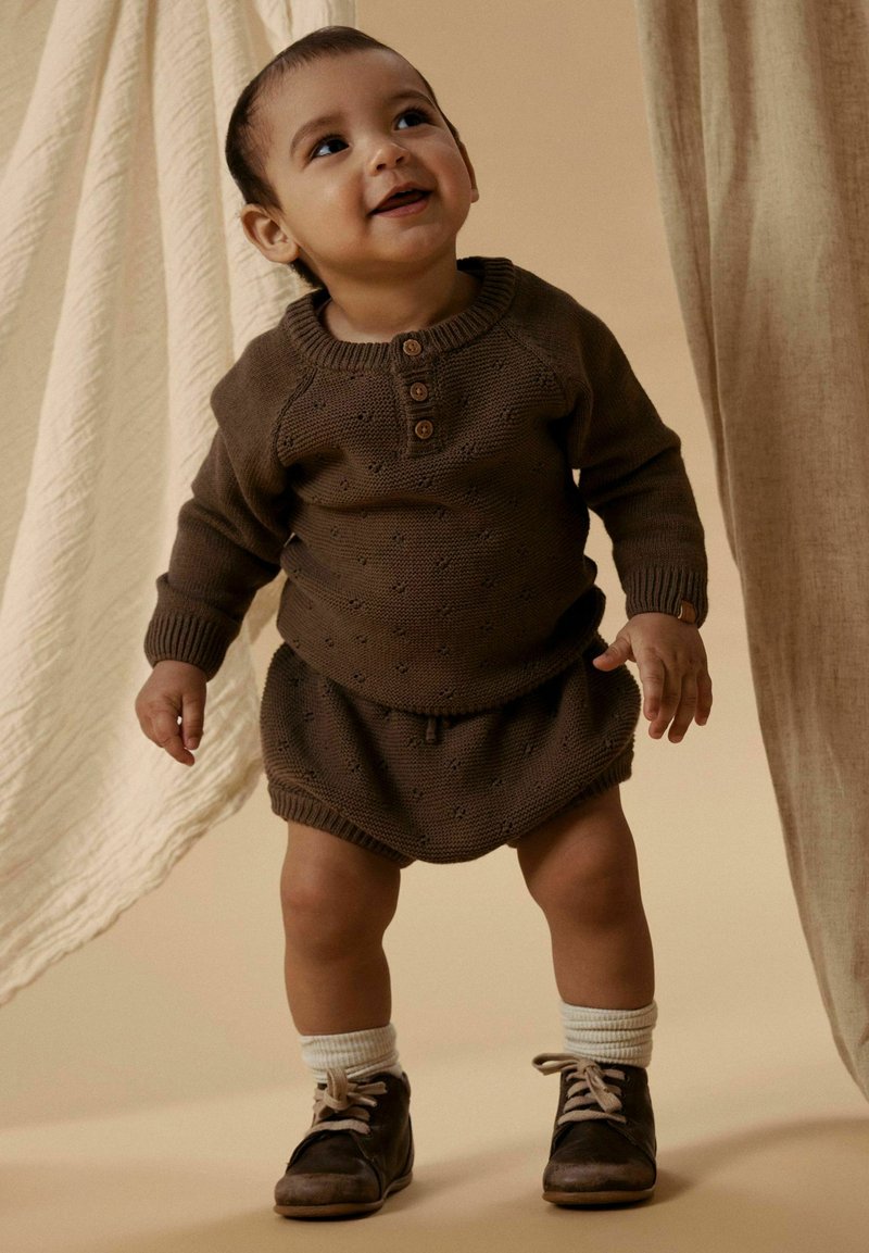 Tout-petit debout et souriant, portant un pull en maille marron et un short, des chaussettes blanches et des chaussures marron foncé, devant un tissu beige drapé.