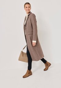 Cappotto lungo in misto lana marrone con due tasche frontali, chiusura a bottoni, abbinato a jeans skinny neri e stivaletti alla caviglia marroni. Borsa beige.