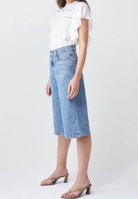 Blouse blanche à volants, culottes en denim bleu taille haute et sandales à talons blocs vertes. Texture lisse et coupe décontractée, longueur au genou.