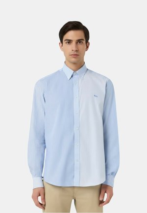 Uomo che indossa una camicia a maniche lunghe con bottoni, caratterizzata da strisce verticali alternate blu e bianche, abbinata a pantaloni beige.