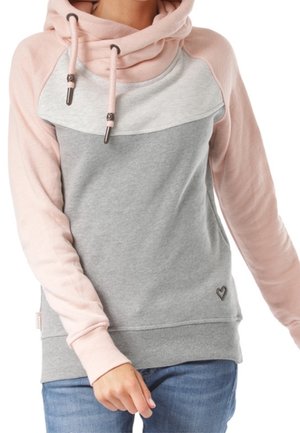Vrouw draagt een hoodie met roze en grijze kleurvlakken en trekkoorden, gecombineerd met een blauwe jeans, zichtbaar van nek tot heupen.