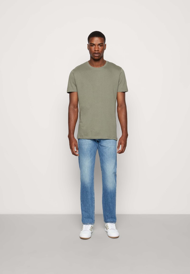 Tricou cu guler rotund de culoare verde măsliniu, din material de bumbac, asortat cu blugi drepti de culoare albastră deschisă și adidași albi cu dungi subtile.