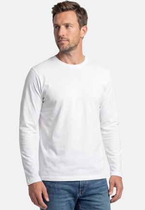 Man met kort bruin haar, gekleed in een eenvoudige witte lange mouwen shirt en blauwe spijkerbroek, die iets naar links kijkt.