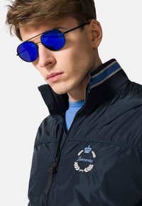 Granatowa kurtka bomberka z wysokim kołnierzem, z haftowanym logo. Model nosi okrągłe niebieskie okulary przeciwsłoneczne i jasnoniebieską koszulkę.